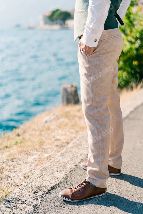 Preview: Man with his hands in his pants pockets stands on the road above the sea. Side view. Cropped