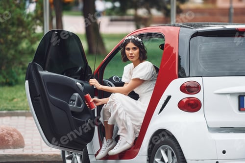 Preview: Sitting in white and red colored car. Bride in dress is outdoors before her wedding begins