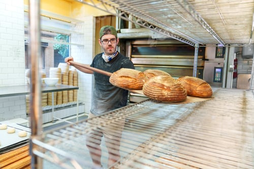 Preview: Artisan Baker Removing Fresh Bread from Oven in Rustic Bakery Setting