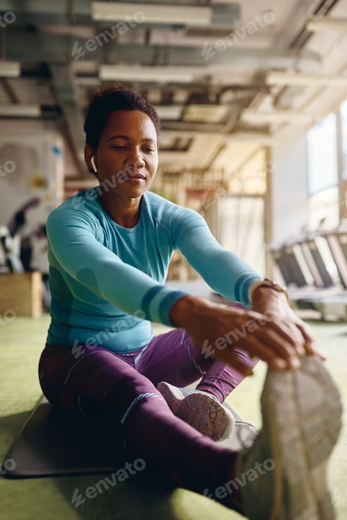 Preview: Black female athlete stretching while warming up for sports training in gym.