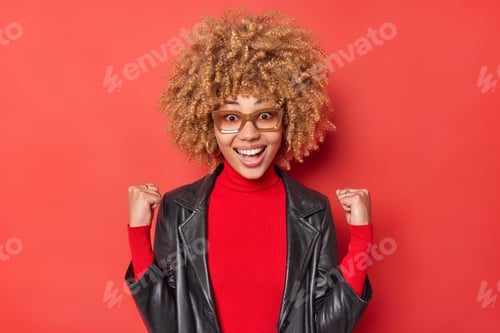 Preview: Positive curly haired woman exlclaims from joy clenches fists with triumph wears turtleneck and leat