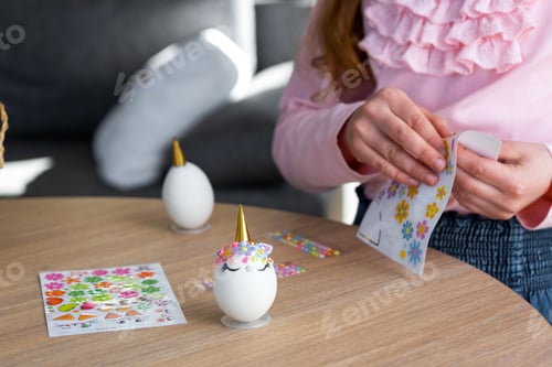 Preview: A cute girl with pink bunny ears makes an Easter craft - decorates an egg in the form of a unicorn w