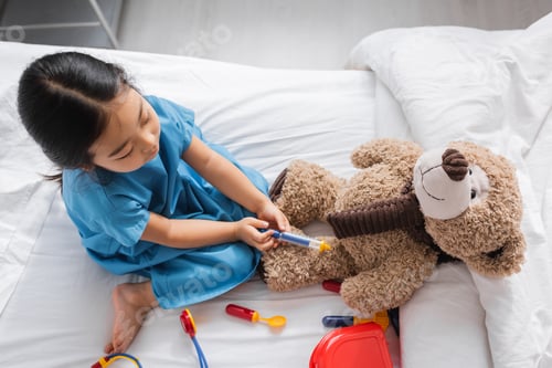 Preview: top view of asian child playing on hospital bed and doing injection to teddy bear with toy syringe