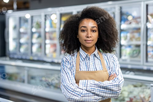 Preview: A confident female worker with crossed arms stands in a grocery store frozen food section, wearing