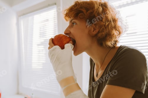 Preview: teenage girl with broken arm in cast in kitchen sitting on countertop eating apple