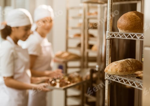 Preview: bakers working at baking manufacture with fresh loaves of bread lying on shelves on foreground