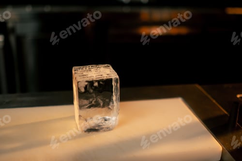 Preview: on the bar counter on white board there is a square cut transparent ice for whiskey
