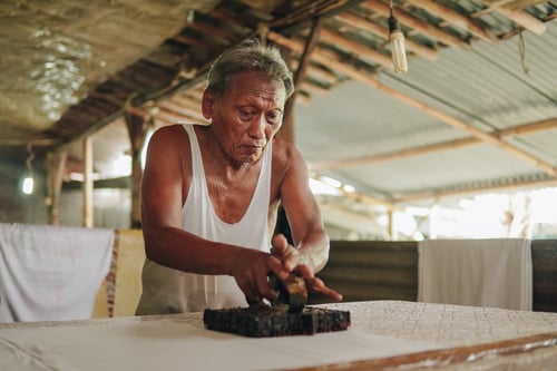 An elderly senior Indonesian man doing the process of stamping batik with a traditional equipment.