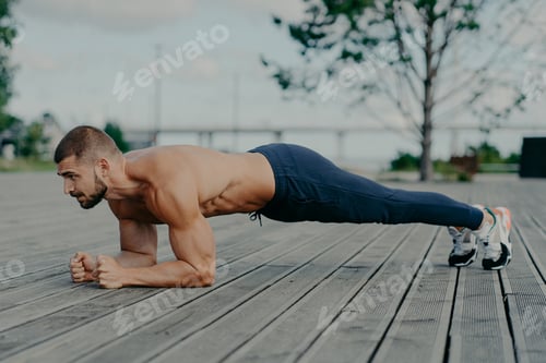 Preview: Motivated male athlete does abs exercise, enjoys bodybuilding training outdoor and stands in plank