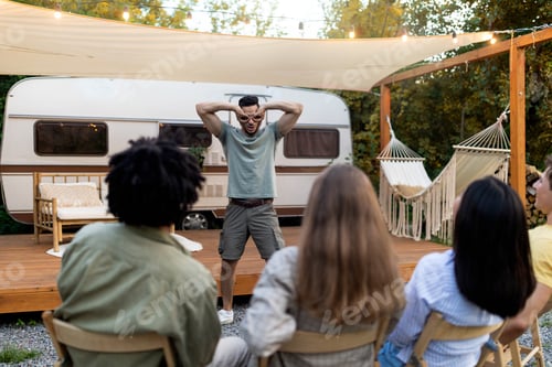 Preview: Group of diverse millennial friends playing charades near trailer, trying to guess word on autumn