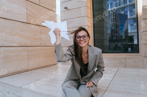Preview: Content businesswoman with glasses sits on steps, playfully waving papers, in a modern cityscape