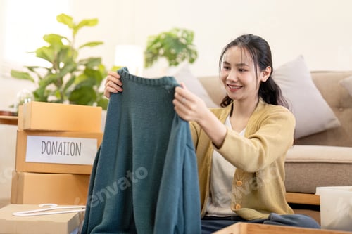 Preview: Joyful woman holding a sweater for donation, organizing clothes for charitable purpose
