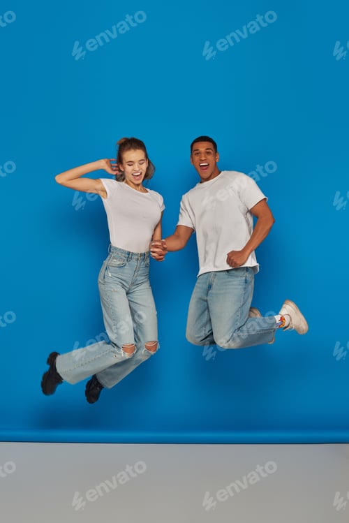 Preview: positive multicultural couple holding hands and jumping on blue background, full length view