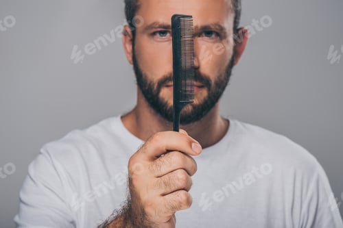 Preview: bearded man holding comb and looking at camera isolated on grey