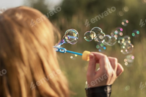 Preview: Red-haired girl blows soap bubbles in the park at sunset . View from the back.