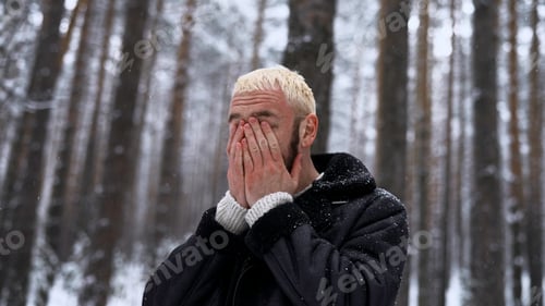 Preview: Stylish hipster man in pine tree winter forest rubbing his cold red face with his hands. Media