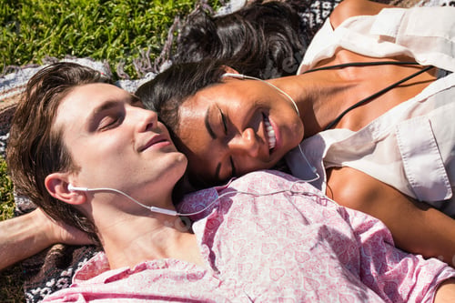 Preview: Young couple relaxing outdoors, lying on blanket on grass, sharing earphones