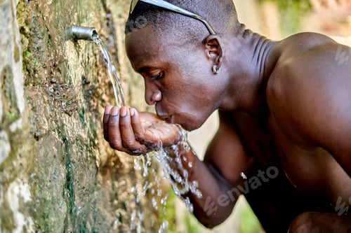 Preview: Shirtless black male traveler sipping clean fresh water from tube on wet stone wall during summer
