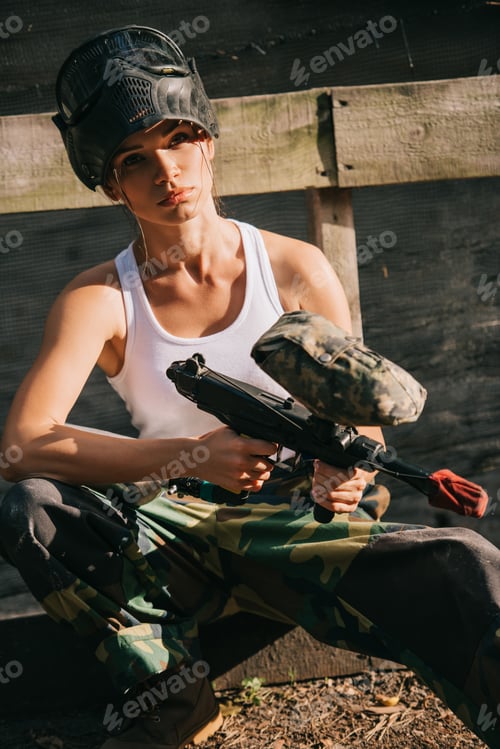 Preview: beautiful young female paintballer in white singlet and goggle mask holding paintball gun outdoors