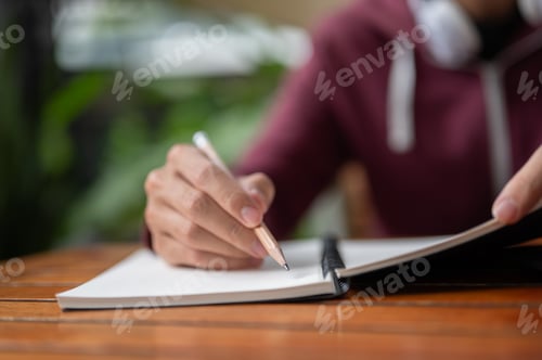 Preview: Close up college student man holding pencil writing or drawing in notebook at wooden table in a cafe