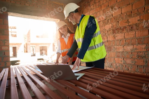 Preview: Foreman and architect in hardhats reviewing paper blueprint on laptop