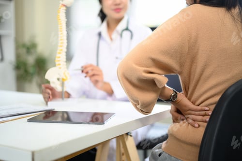 Preview: Patient holding lower back in pain while consulting doctor with spine model in clinic