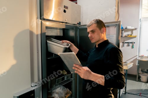 Preview: chef stands in kitchen in a black uniform and looks through what is on the list in the refrigerator
