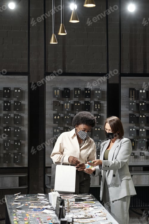 Preview: Young shop assistant in protective mask showing necklace to female consumer