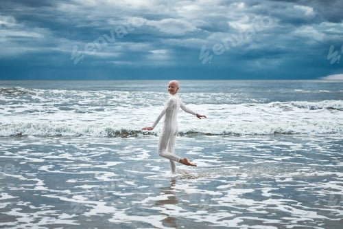 Preview: Happy hairless girl with alopecia in white futuristic suit dancing on beach bathed by ocean waves