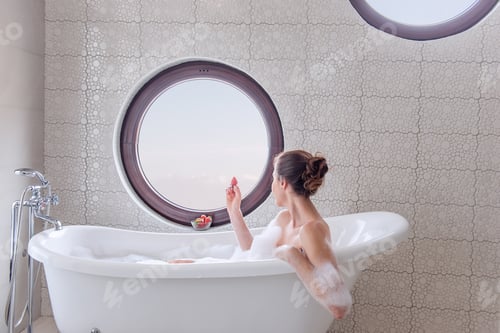 Preview: Young woman smilling in white, modern bath with foam eats juicy strawberry.