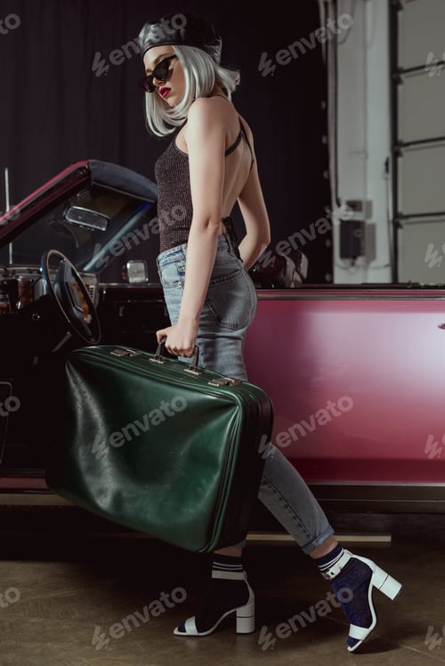 Preview: full length view of stylish blonde woman in sunglasses and beret holding old-fashioned suitcase bear