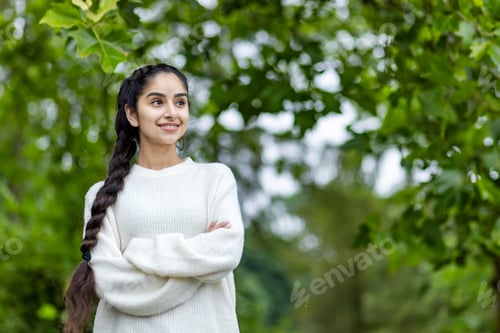 Preview: A happy and smiling Indian woman in a white sweater and a braided hairstyle stands in the park with