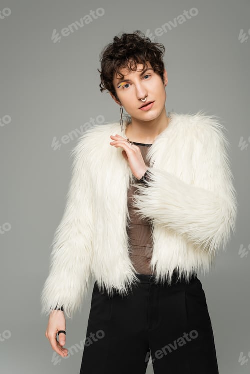 Preview: fashionable nonbinary person in white faux fur jacket looking at camera isolated on grey
