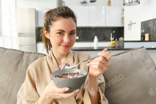Preview: Close up portrait of cute woman on sofa, eating breakfast, holding delicious chocolate cereals with