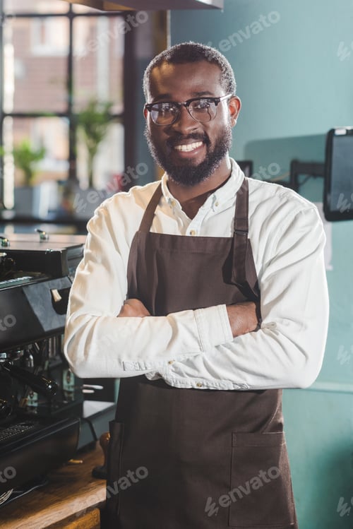 Preview: happy young african american barista standing with crossed arms and smiling at camera in coffee shop