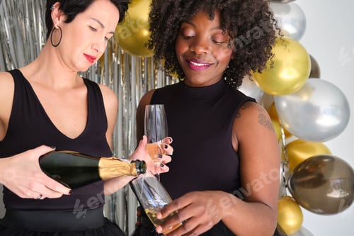 Preview: Two women serving champagne glasses from bottle and celebrating New Year in a party.