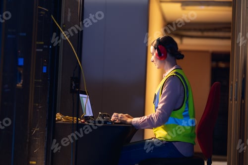 Preview: Caucasian female engineer wearing ear plugs using a laptop in computer server room