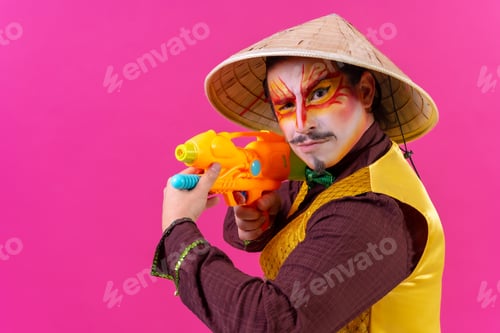 Preview: Clown with white facial makeup on a pink background, having fun with toy guns