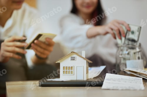 Preview: A house model and bills on table with couple planning to buy their future new house on background