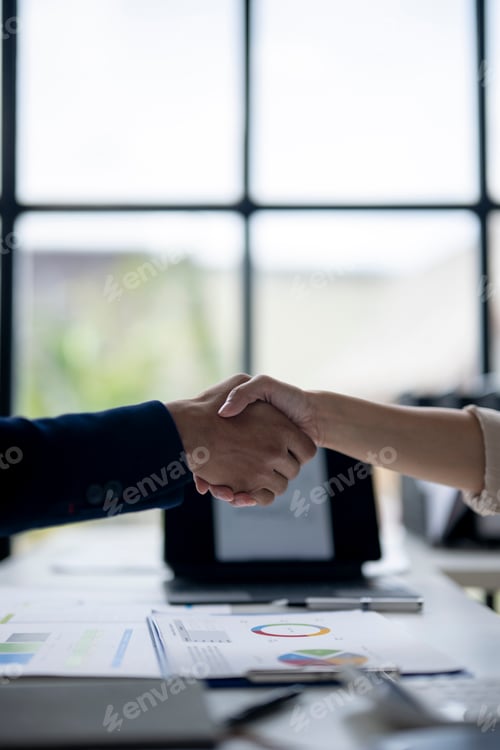 Preview: Two people shaking hands in front of a table with a laptop and papers
