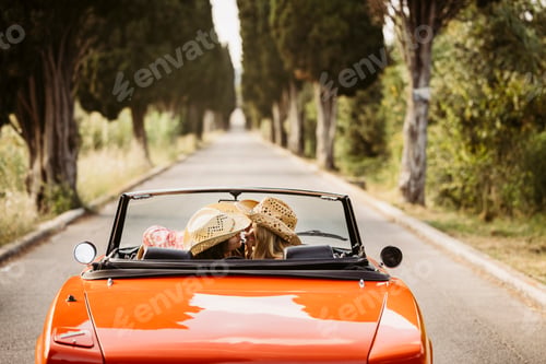 Preview: Two women kissing while driving orange convertible car on a country road