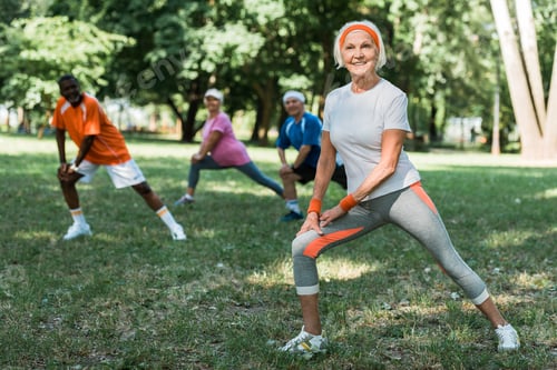Preview: selective focus of cheerful retired woman stretching near multicultural pensioners on grass