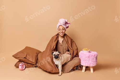 Preview: Indoor shot of positive woman stays in bed under blanket with pug dog plays with favorite pet wears