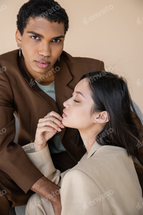 Preview: stylish african american guy with piercing looking at camera near young asian woman with closed eyes
