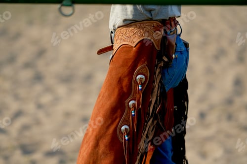 Preview: Close-up of a cowboy wearing a leather chaps and denim jeans standing in a sandy arena.