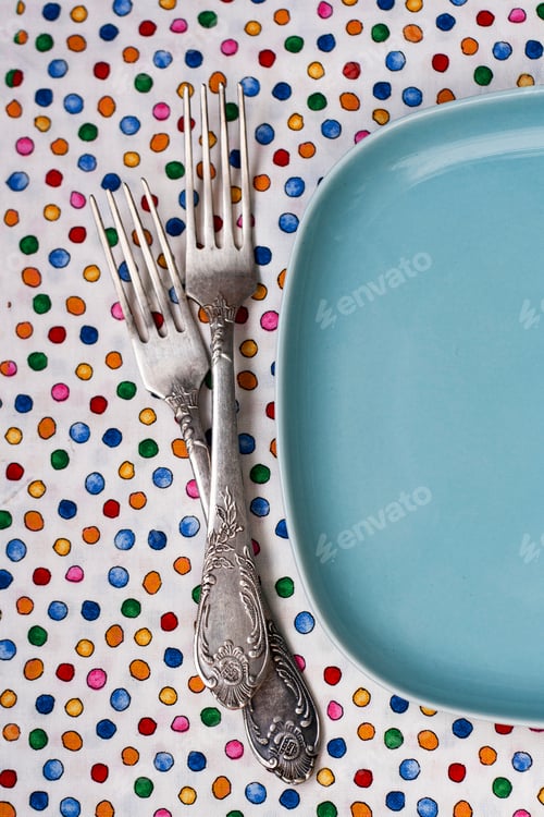 Preview: Two forks lie on a tablecloth next to a blue plate.