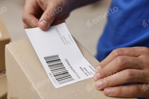 Preview: Man sticking shipping label with barcode on parcel indoors, closeup