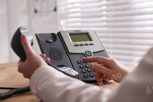 Preview: Woman using desktop telephone at wooden table in office, closeup. Hotline service