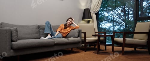 Preview: Young woman relaxing on a sofa in cozy living room with modern decor, wearing stylish clothes and