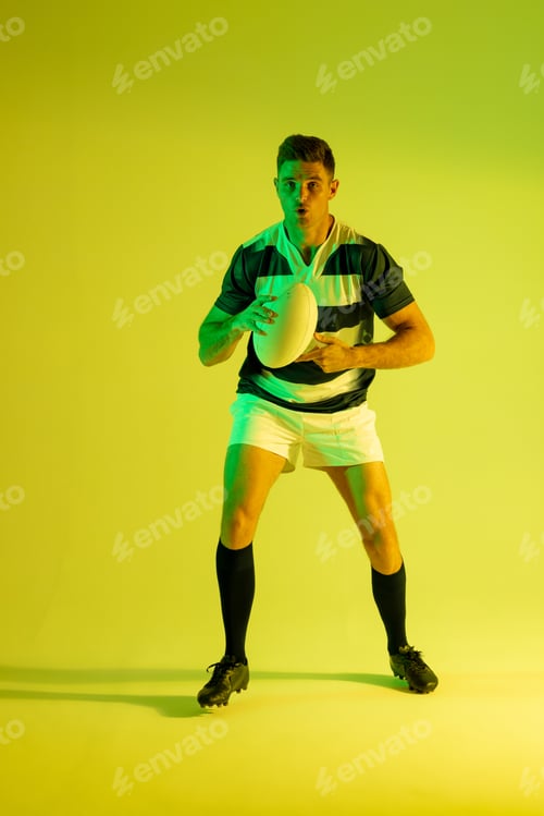 Preview: Caucasian male rugby player with rugby ball over yellow lighting